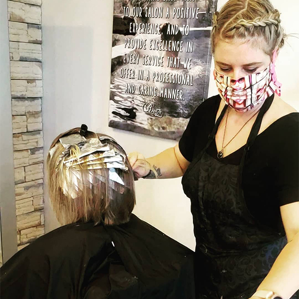 A hairstylist applying foil highlights to a client's hair in a Texas salon, wearing a face mask and standing near a wall plaque.