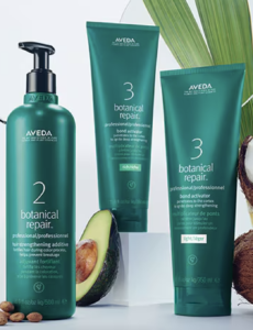 Three Aveda Botanical Repair hair care products, including a pump bottle labeled 2 and two tubes labeled 3, are displayed with avocado and coconut ingredients in the background, showcasing the health-focused power of this treatment lineup.